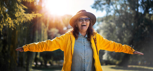 Beautiful happy senior mature woman in hat and yellow jacket enjoying nature in the park stretching...