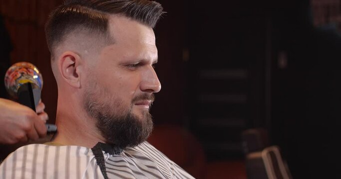 Close-up of a handsome Caucasian guy sitting in a hairdressing salon. An unrecognizable barber shaves a bearded man's head with a trimmer. 4K 50 fps slow motion 