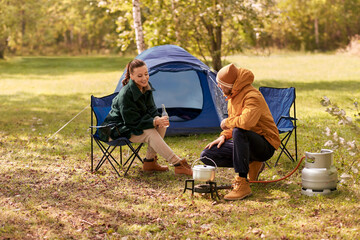 camping, tourism and travel concept - happy couple drinking beer and cooking food in pot on tourist...