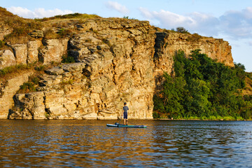 A man on a SUP on the river in summer at sunset against the backdrop of a rock