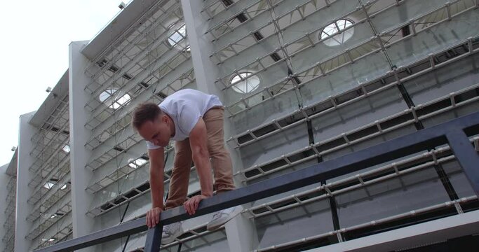 Professional acrobat performing dangerous trick on the railings against backdrop of modern stadium. Caucasian guy  making a handstand with straddle split on a narrow metal pipe
50 fps slow motion
