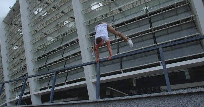 Professional acrobat performing dangerous trick on the railings against backdrop of modern stadium. Caucasian guy  making a handstand with straddle split on a narrow metal pipe
50 fps slow motion
