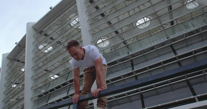 Professional acrobat performing dangerous trick on the railings against backdrop of modern stadium. Caucasian guy  making a handstand with straddle split on a narrow metal pipe
50 fps slow motion
