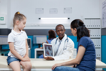 Obraz premium African american pediatrician doctor holding tablet computer with heart radiography on screen discussing xray medical expertise with mother during examination.Therapist man working in hospital office
