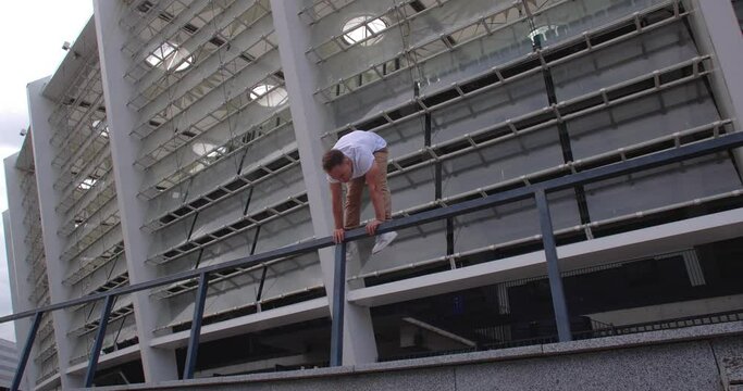 Professional acrobat performing dangerous trick on the railings against backdrop of modern stadium. Caucasian guy  making a handstand with straddle split on a narrow metal pipe
50 fps slow motion

