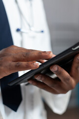 Closeup of african american doctor holding tablet computer typing healthcare treatment after analyzing sickness expertise during clinical appointment. Therapist man working in hospital office