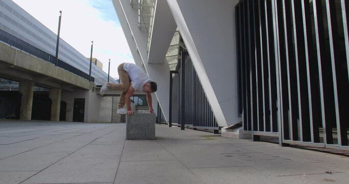 Professional acrobat performing trick on the street against backdrop of modern stadium. Caucasian guy  making a handstand with straddle split on a playground  50 fps slow motion
