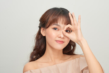 Beautiful young Asian woman show OK sign over her eye on gray background