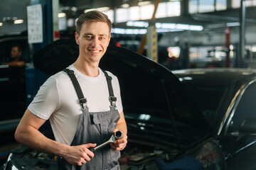 Medium shot portrait of cheerful handsome young mechanic male wearing uniform holding special key ratchet wrench, in auto repair shop garage with vehicle background, selective focus.
