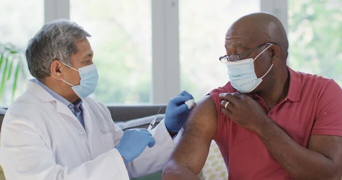 Male doctor wearing face mask vaccinating african american senior man at home