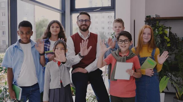 Male teacher with middle school children looking at camera and waving hands in modern school. Happy middle school childen. High quality 4k footage