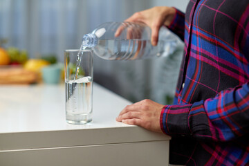 Frame image of a female hand holding drinking water bottle and pouring water into glass on table on kitchen background.