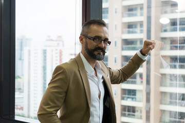 Bearded businessman executive in suit looking at camera using smartphone on window with city building background.