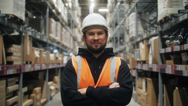 Portrait of worker warehouse wearing vest and safety helmet standing in retail warehouse full of shelves with boxes.