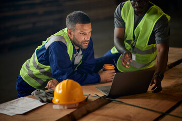 Warehouse workers read delivery plans on laptop while working at lumber storage compartment.