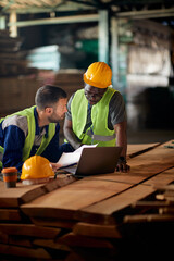 Two male workers cooperate while analyzing paperwork and using laptop at distribution warehouse.