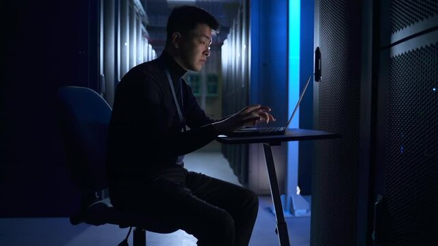 Man engineer working with computer and sitting at table in dark database corridor spbas. Young male specialist types text and looks at laptop screen, does analytical job and sits at desk in interior