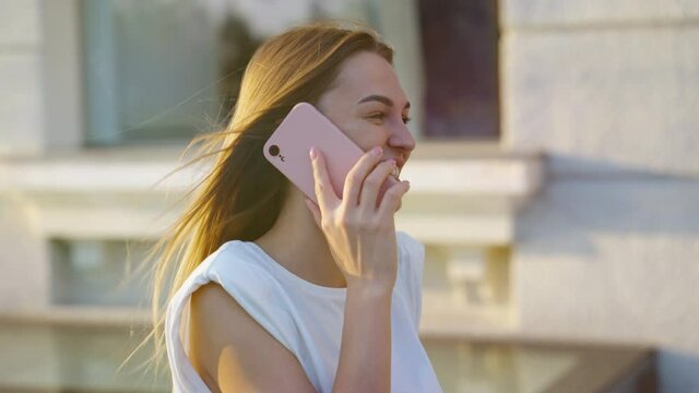 Beautiful woman with long blonde hair wearing white top talking on phone while walking on street, camera rotation. parallax effect. Happy female in rays of summer sunset. Concept of communication