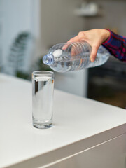 Closeup of a Ffemale hand holding drinking water bottle and pouring water into glass on table on kitchen background.