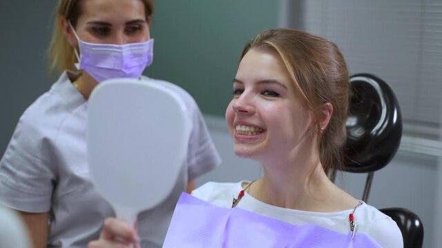 Young woman patient looking at teeth and smiling, doctor standing nearby in clinic office spbas. Close-up view of beautiful female holds mirror and looks with smile at aligners in her mouth