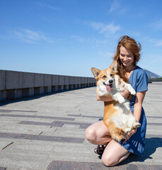 A girl with long red hair in a blue dress hugs her corgi dog in the park, in the afternoon. Friendship, vaccination of dogs, morning walk, rest, domestication