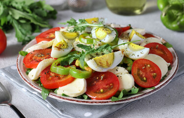 Healthy salad with tomatoes, mozzarella, olives, arugula and eggs on a plate on a light gray background.