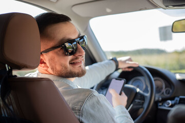 Portrait of young male driver using his mobile phone while driving the car and smiling. Man driver using smart phone in car.