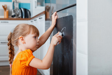 Little caucasian focused child girl draw with chalk on chalk board on refrigerator in kitchen at...