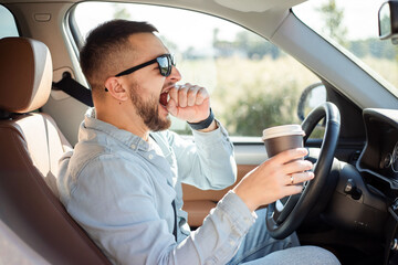 Young handsome man feeling tired and yawning while driving a car.