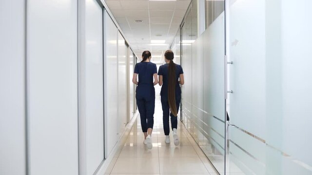 Two female doctors in medical blue suits with long beautiful hair walk in the corridor of a modern clinic discussing the medical history of a patient with heart failure work issues.