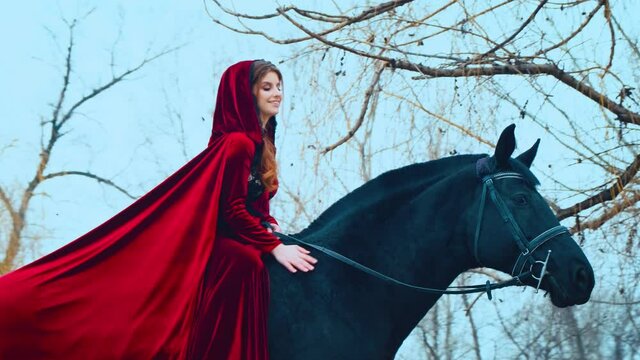 Medieval woman princess in red dress sits astride black steed horse. Girl rider in vintage hood, cloak velvet cape train flies in wind. Background autumn trees forest, blue sky spring winter nature
