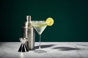 Martini glass with cocktail or mocktail and lime wedge, shaker and jigger on dark background