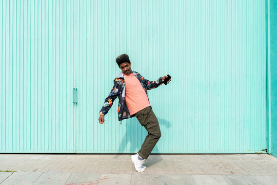 Ecstatic man holding smart phone while dancing in front of turquoise wall