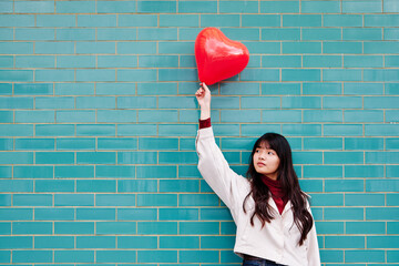 Young woman looking away while holding red heart shape balloon in front of brick wall