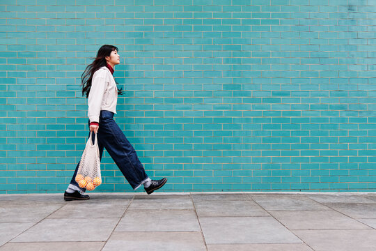 Young woman with mesh bag on footpath by turquoise brick wall