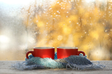 two red cups wrapped together in a blue scarf against the background of a wet window after the rain. warming autumn coffee