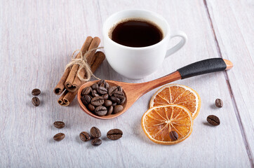 A cup of fresh Americano, a wooden spoon with coffee beans, cinnamon sticks and dried oranges on a light wooden background.