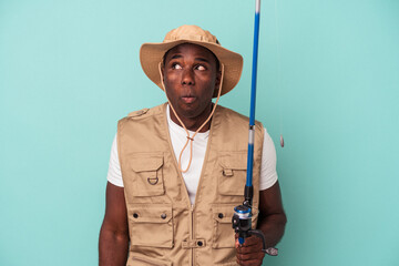 Young African American fisherman holding rod isolated on blue background shrugs shoulders and open eyes confused.