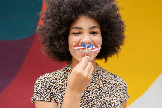 Afro woman holding mustache prop in front of wall