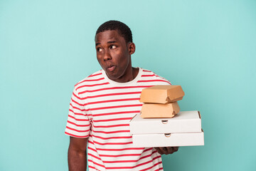 Young African American man holding pizzas and burgers isolated on blue background shrugs shoulders...
