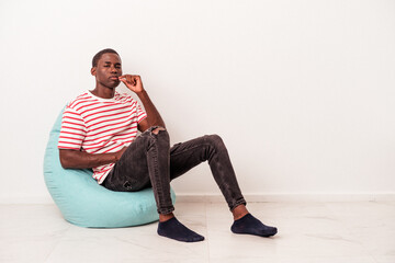 Young African American man sitting on a puff isolated on white background with fingers on lips keeping a secret.