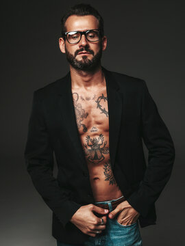 Portrait of handsome confident stylish hipster lambersexual model. Sexy modern man dressed in black jacket. Naked torso with tattoos.Fashion male posing in studio on dark background. In spectacles
