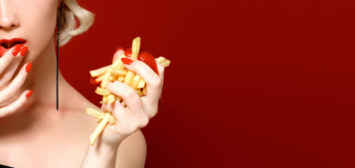 Unhealthy eating. Junk food concept. Portrait of fashionable young woman holding eating fried potato fries, chips in her hand and posing over red background. Close up. Copy space. Studio shot