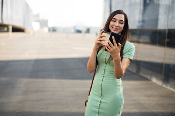 Fototapeta premium Young girl with coffee to go holding smartphone. Beautiful woman talking to the phone while enjoying in fresh coffee