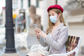 Asian beautiful woman wearing suit and red cap holds smartphone in her hands while sits on bench in park while wearing medical face mask in new normal lifestyle concept