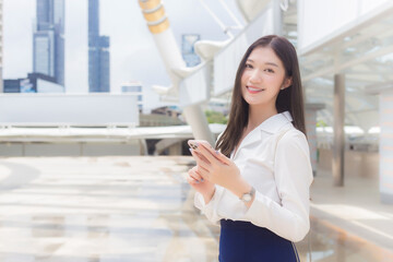 Beautiful young Asian working woman is holding smartphone standing outdoors and smiling happily while she go to the office in big city with business buildings and city in background.