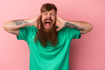 Young caucasian ginger man with long beard isolated on pink background covering ears with hands trying not to hear too loud sound.