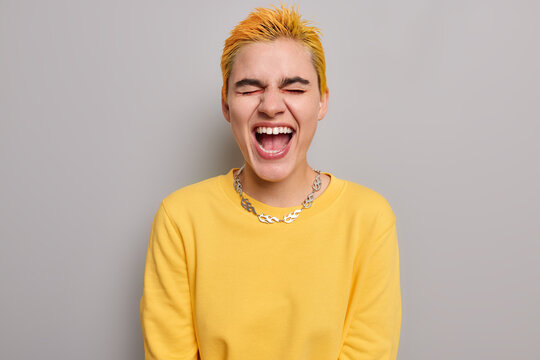 Photo of overjoyed hipster girl exclaims loudly keeps mouth opened has short dyed hair closes eyes and shouts wears casual yellow jumper with metal chain around isolated over grey background