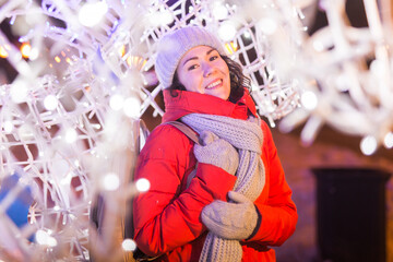Girl having fun on christmas decoration lights street. Young happy smiling woman wearing stylish knitted scarf and jacket outdoors. Model laughing. Winter wonderland city scene, New Year party.