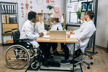 Muslim scientist doctor in hijab holding laptop in hands and looking at her colleagues during conference in modern medical center. Brainstorming of medical staff at boardroom.